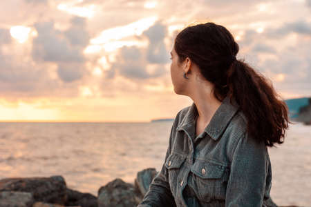 A young woman sits alone by the sea and looks at the sunset. View from the back. The concept of psychology and mental health.の写真素材