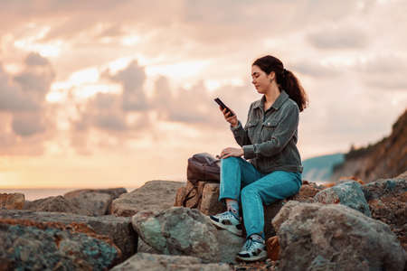 A young woman is sitting on rocky rocks and using a phone. Sunset in the background. Communication concept.の写真素材