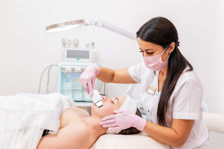 Beauty salon. A cosmetologist wearing a medical mask and rubber gloves cleans the face using an ultrasound machine. In the background is a cosmetic device. Concept of professional cosmetology and beauty.の写真素材