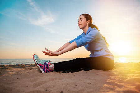 Young caucasian woman in sportswear sitting on the sand and performs a stretching of the legs. In the background of the sunset and the sea.の写真素材