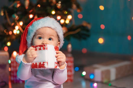 Portrait of cute baby in a Christmas hat holding a holidays gift. Festive lights and xmas gifts on the background. Merry Chrismas and Happy New Year concept.の写真素材