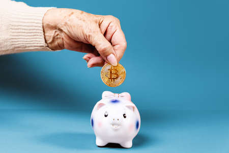Wrinkled hand of an elderly woman puts a bitcoin in a piggy bank. Blue background. The concept of...