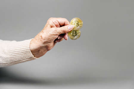 Wrinkled hand of an elderly woman holding a two golden bitcoin. Gray background. Copy space. The concept of online investment and cryptocurrency.の写真素材