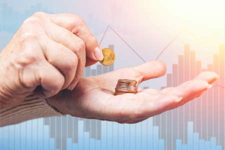 Close up of females wrinkled hands putting a euro coin in a stack of coins. Business chart on the...
