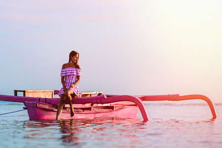girl sitting in a pink boat in the middle of the ocean at sunset.の写真素材