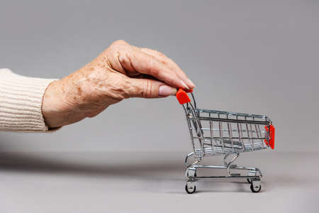 Hand of elderly woman rolls an empty little shopping cart. Gray background. Concept of expenses and poverty.の写真素材
