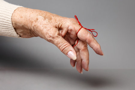 Hand of a senior woman with a red thread tied on her index finger. Gray background. The concept of Alzheimer's and Parkinson's diseases.の写真素材