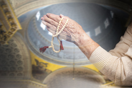 Wrinkled hands of a senior woman folded in prayer and holding a rosary. The dome of the church in the background. Double exposure. The concept of religion.の写真素材
