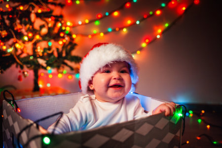 Christmas. Portrait of a happy baby in a Christmas hat sitting in a gift box. A garland lights up in the background. The concept of winter Christmas holidays.の写真素材