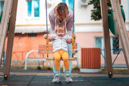 A young mother rides her little daughter on a swing on the playground. The concept of motherhood.の写真素材
