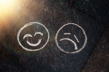 Smiling and sad emoji drawn with crayons on the ground. Rainy and sunny weather. The concept of psychological and mental health.の写真素材