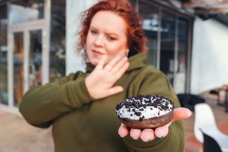 A blurry portrait of a well-fed woman holding a chocolate donut in the palm of her hand. The concept of diet and weight loss.の写真素材