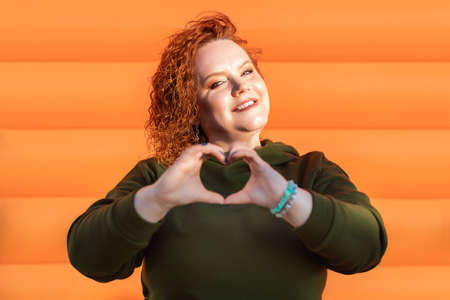 Portrait of a young woman showing a heart gesture with her hands. Orange striped background. The concept of social networks and psychology.の写真素材