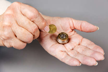 Close up of females wrinkled hands putting a euro coin in a stack of coins. Gray background. The concept of pension savings and retirement.の写真素材