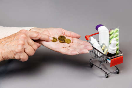 Poverty. Hands of elderly woman counting a coinson near full of medicines little shopping cart. Gray background. Concept of online shopping.の写真素材