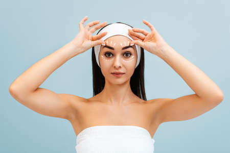 Portrait of woman in a headband and a bath towel pulls her eyebrows with her hands. White markings above the forehead. The concept of massage and plastic surgery.の写真素材