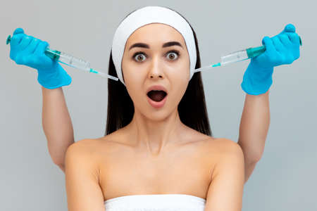 Portrait of a young frightened woman, behind which a cosmetologist holds two syringes. Gray background. The concept of fear of plastic surgery.の写真素材