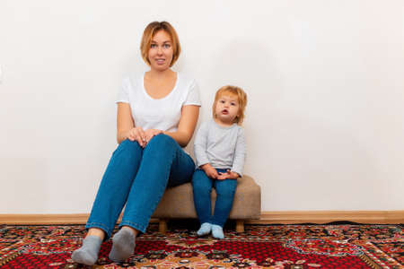 A young mother and her little child are sitting on an ottoman in their new home. The concept of mortgage and purchase of new housing.の写真素材