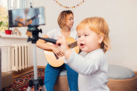 A young mother playing the guitar and records a live video broadcast on a smartphone. A child in a blur in the foreground. The concept of video streaming and online learning.の写真素材