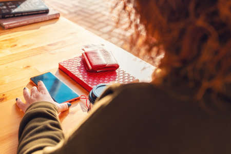 A woman uses a smartphone while sitting at a table. Wallet with money and a notebook lie near on the...