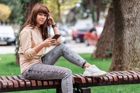 Pretty girl sits on a park bench and holding a smartphone. Outdoor. Free wi-fi area.