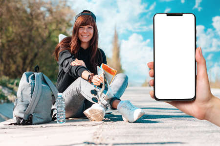 Hitchhiking. Young smiling woman in cap sitting along the road with back pack and paper map. Female hands holding a smartphone with mock up. Local travel and gps concept.の写真素材