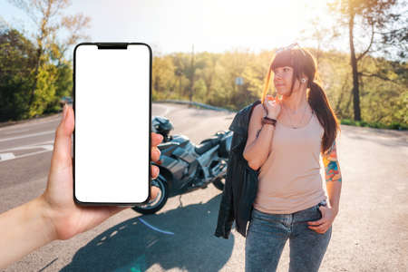 World Motorcyclist Day. A young woman posing against the background of a motorcycle by the road. Female hand holding a smartphone with mock up. Motorcycle travel concept.の写真素材