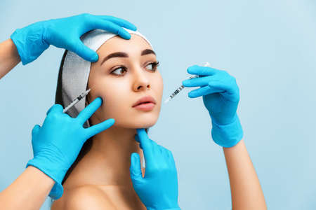 Portrait of a beautiful young woman with hands of cosmetologists in medical gloves lean syringes...