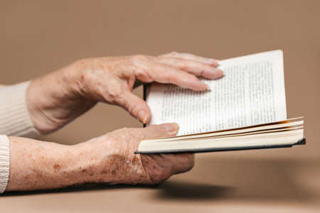 Wrinkled hands of a senior woman are holding a book. Beige background. The concept of education and readingの写真素材