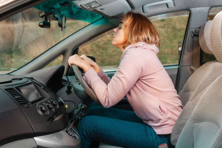 A young blonde woman is sitting at the wheel of a right-handed car and nervously looks at the road....