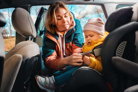 A young caucasian mother prepares her small cute child for a trip by car. A woman fastens her seat...