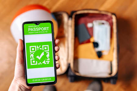 The concept of new normal. Close up view of a male hand holds a smartphone with an immune digital health passport. Top view. A suitcase with clothes and protective masks on the blurred background.の写真素材