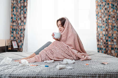 The concept of diseases and seasonal colds. A young brunette woman is sitting on the bed, wrapped in...