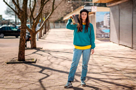 A young Caucasian hipster woman poses with a skateboard over her shoulders. In the background, an alley. Concept of sports lifestyle and street culture.の写真素材