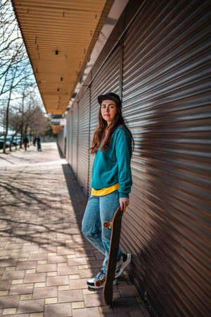 Young beautiful hipster woman posing with her hand on a skateboard. In the background, a street with...