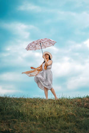 Concept of psychology and loneliss. A young woman in a straw hat and dress, holding an umbrella blown away by the wind. Cloudy sky in the background.の写真素材