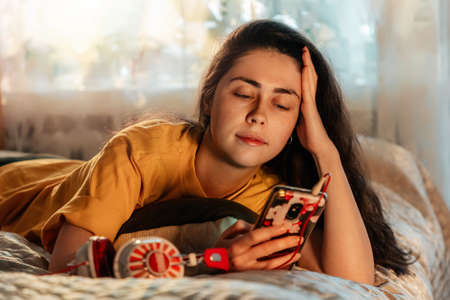 Portrait of smiling young woman is lying on bed and listening to music, putting headphones to her ear, looking into her smartphone.の写真素材