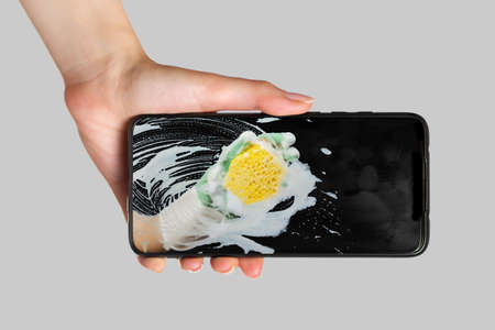 A female's hand is holding a smartphone. On the phone screen, a hand in rubber gloves cleans dirt with a washcloth and foam. The concept of online ordering a professional cleaning service.の写真素材
