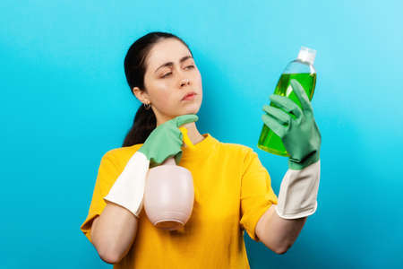 Portrait of a young thoughtful woman in rubber gloves reading the composition on a bottle of household chemicals. blue background. The concept of eco-friendly cleaning.の写真素材