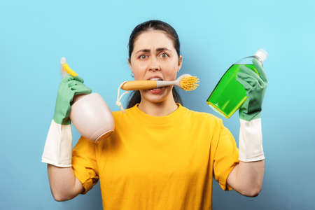 Portrait of a stressed Caucasian young woman in rubber gloves, holding a brush in her mouth and household cleaning products in her hands. blue background. Cleaning day concept.の写真素材