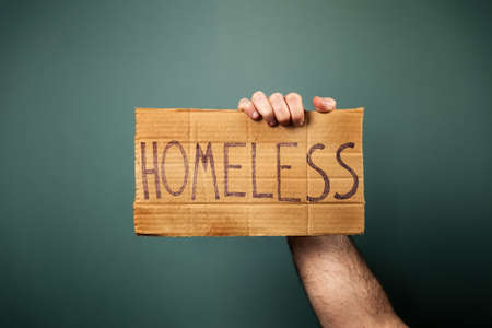 A male's hand holds a cardboard sign with the inscription homeless. Dark background of a greenish...