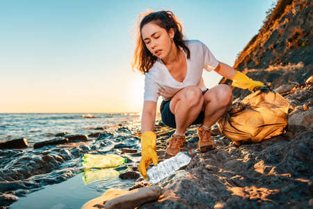 A young woman volunteer squats and picks up garbage on the ocean shore. Cleaning of the coastal zone. Sunset in the background.の写真素材