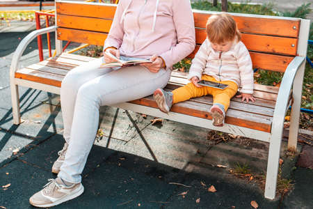 Mother and her little child sitting on a bench and relaxing with book and smartphone. The concept of a current family relaxation in the park.の写真素材
