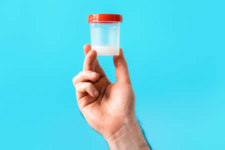 A male's hand is holding a plastic jar with a sperm analysis in close-up. blue background. The concept of a sperm bank and artificial insemination.の写真素材