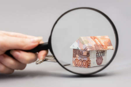 Female hand holding a magnifier glass and showing zoomed at paper origami from russian rubles. gray background. Concept of mortgage and leasing of real estate.の写真素材