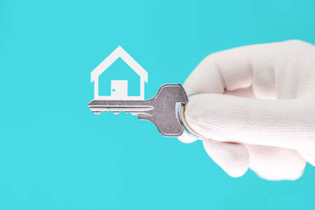 Close-up of a hand in a white glove holding a new metal key on a bright turquoise background. The symbol of the house. Leasing and mortgage concept.の写真素材