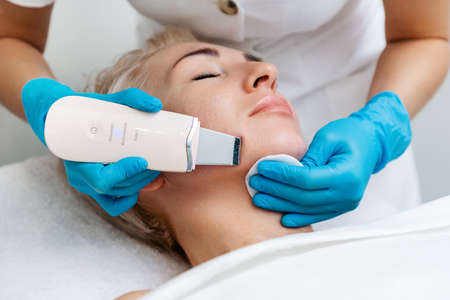 A beautician in blue gloves does an ultrasound cleaning of a woman's face. Cleansing and rejuvenating procedure in a beauty salon.の写真素材