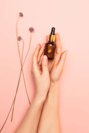 Beautiful female hands hold a brown glass bottle with care cosmetics. Pink background with dry flowers. flat lay. vertical. Concept of beauty products.の写真素材