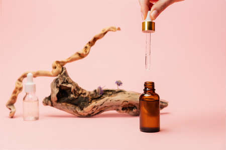 A woman drips facial serum from a pipette into bottle. Close-up of hands on a pink background with cosmetic and natural decor. The concept of beauty care products.の写真素材