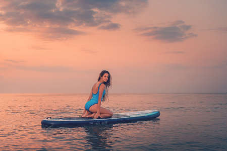 A young sexy tanned woman poses sitting on a sup board. back view. Sunset in the background. copyspace. Concept of sport and surfing.の写真素材